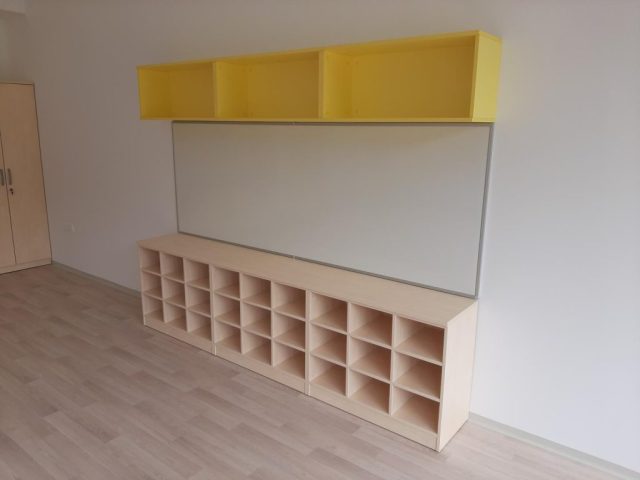 Regal z odprtimi predali in rumenimi zgornjimi omaricami v šolski učilnici